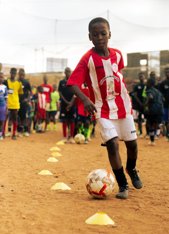 A boy in red soccer jersey focuses on a ball in an outdoor training session.